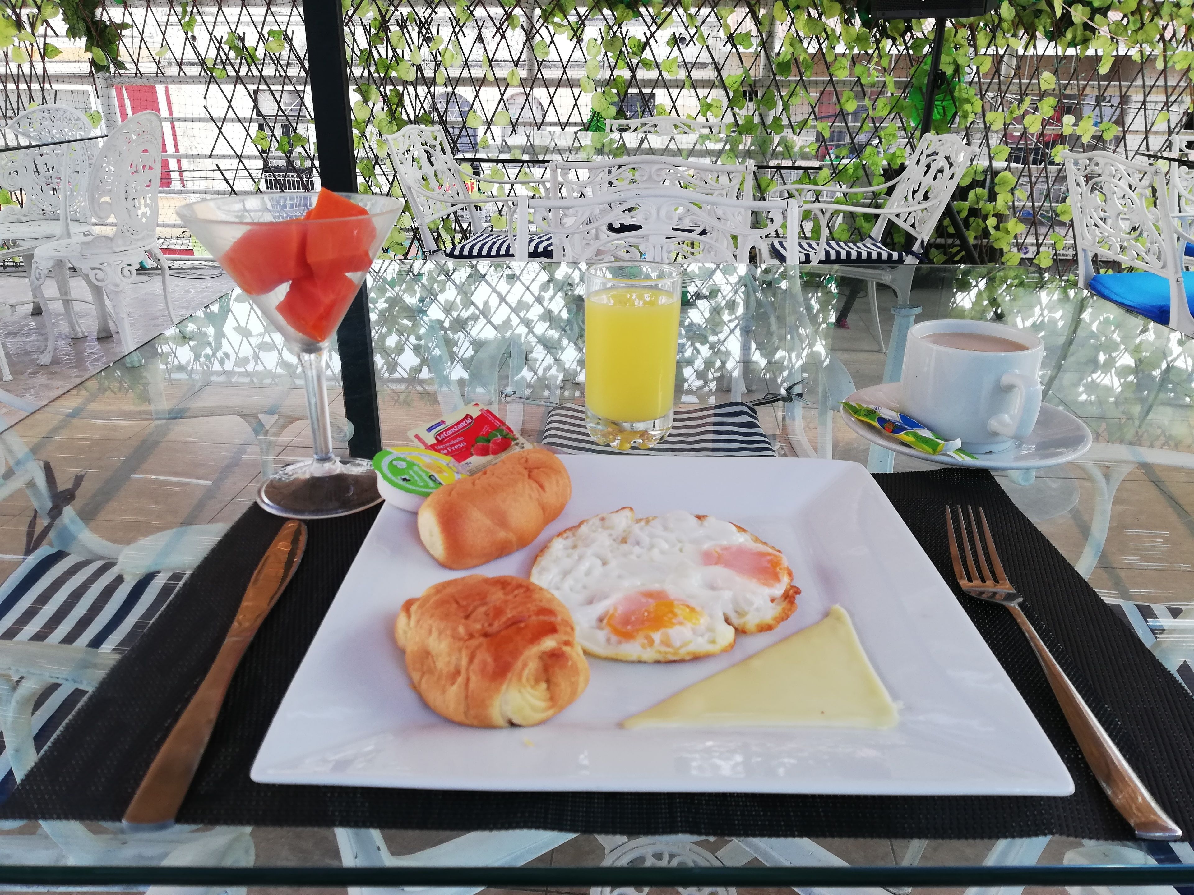 Une table en verre est dressée pour le petit-déjeuner : on y trouve une assiette blanche, un verre de jus d’orange, une tasse de café et une fourchette.