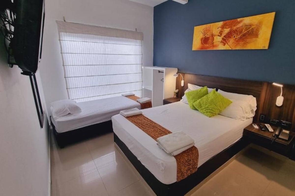 A hotel room with a bed, a small desk, a window with blinds, a nightstand with a lamp, and a painting on the wall.
