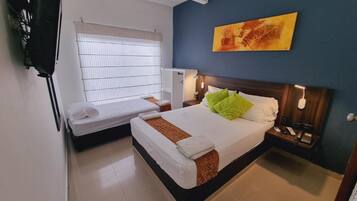 Una habitación de hotel con una cama, un escritorio pequeño, una ventana con persianas, una mesita de noche con una lámpara y un cuadro en la pared.
