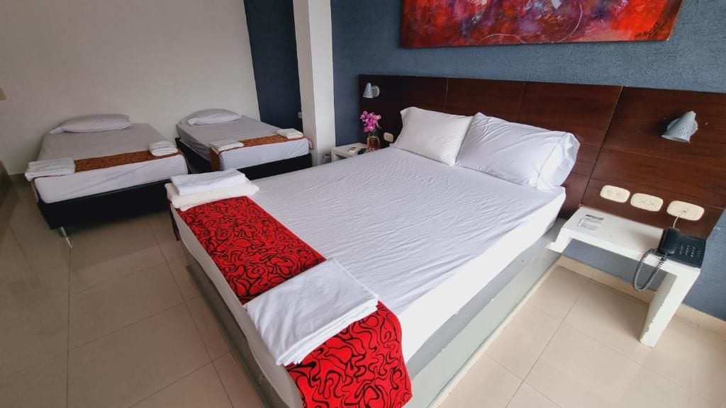 A hotel room with a large bed, two smaller beds, a bedside table with a lamp, and a painting on the wall.