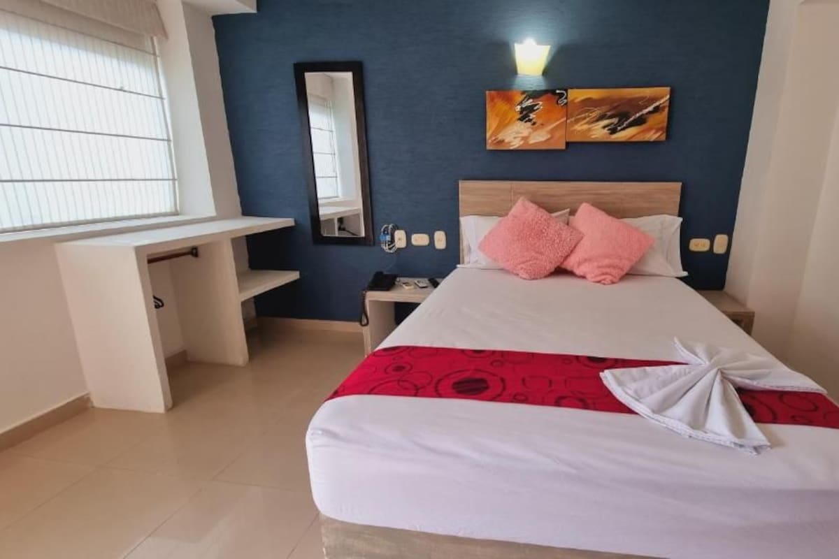 A hotel room with a bed, a desk, a chair, a nightstand, and a wall-mounted artwork.