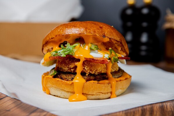 A sandwich with melted cheese, bacon, and greens on a bun, placed on a white paper on a wooden surface. - OYO Weaste Hotel (Salford)
