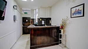 A reception desk with a wooden finish, a white tiled floor, a framed picture on the wall, and a small potted plant. - OYO Weaste Hotel (Salford)