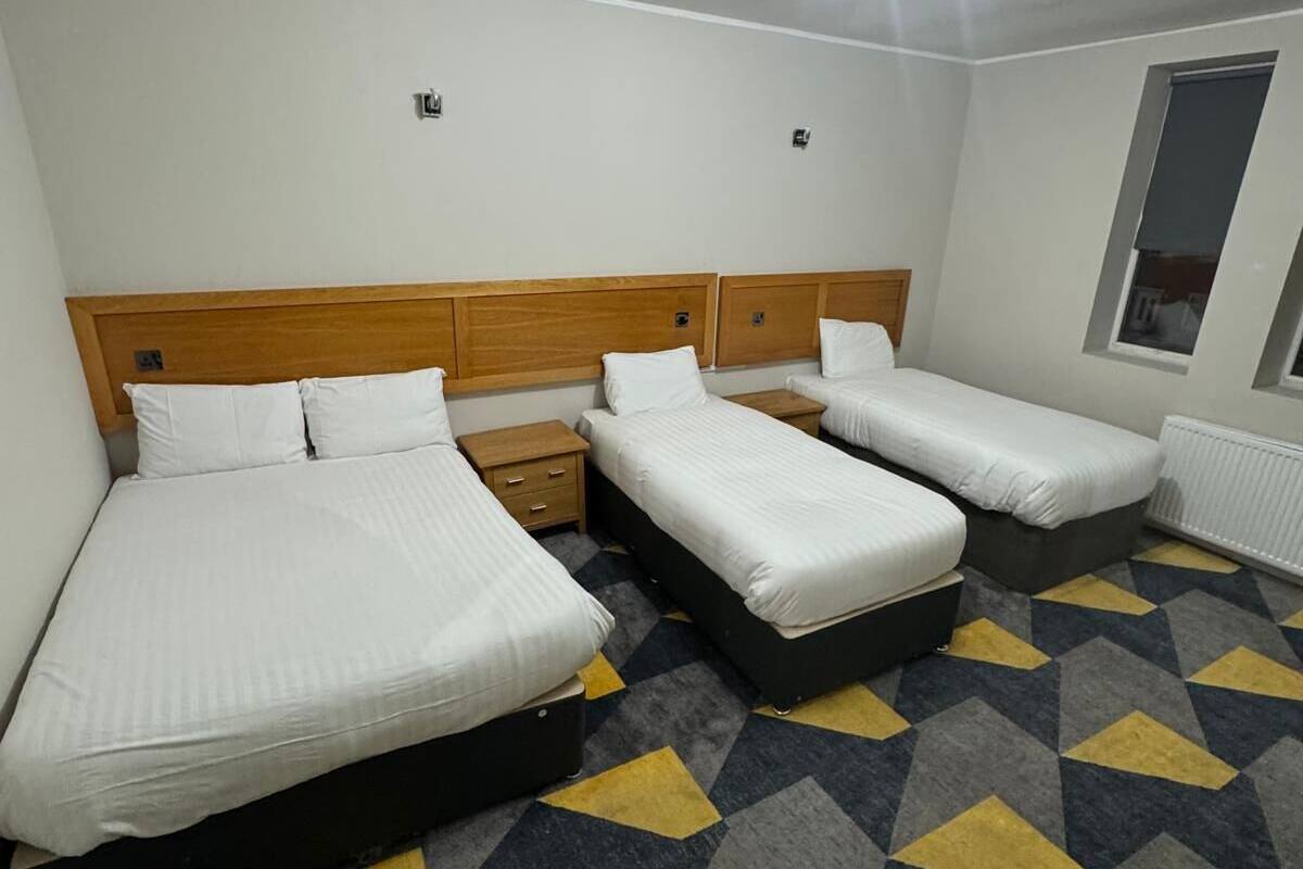 Une chambre d’hôtel avec un grand lit, deux petits lits, une table de chevet, une commode, un radiateur et une moquette à motifs.