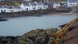 A coastal town with white buildings, rocky shoreline, and a red buoy in the water. - The Old School (Islay Island)