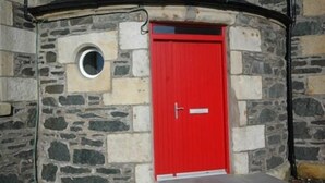 A stone building with a red door and a small circular window. - The Old School (Islay Island)
