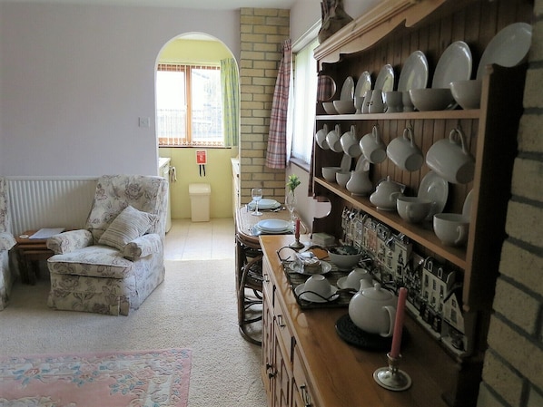 A cozy living room with a floral armchair, a wooden sideboard with dishes, and a view through an arched doorway. - Cae y Rhedyn Annexe (Crickhowell)