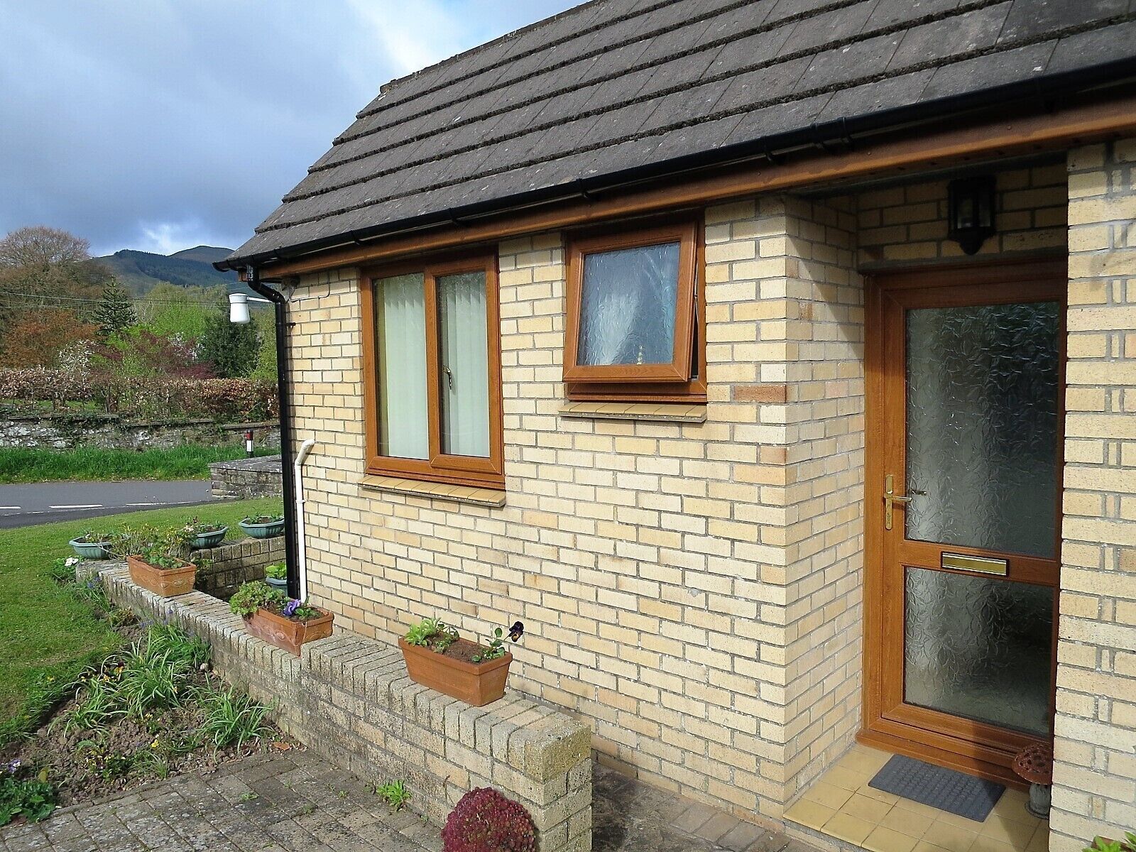 A single-story brick house with a tiled roof, a front door with a glass panel, and a small porch with potted plants.