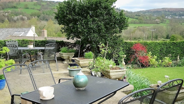 Un patio al aire libre con sillas y una mesa de metal, rodeado de plantas en macetas y vistas a un paisaje verde.