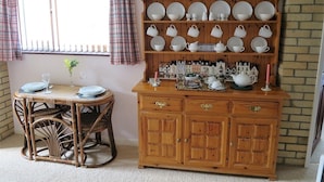 Un comedor con un aparador de madera que exhibe platos y tazas, una mesa de comedor de mimbre con sillas y una ventana con cortinas a cuadros.