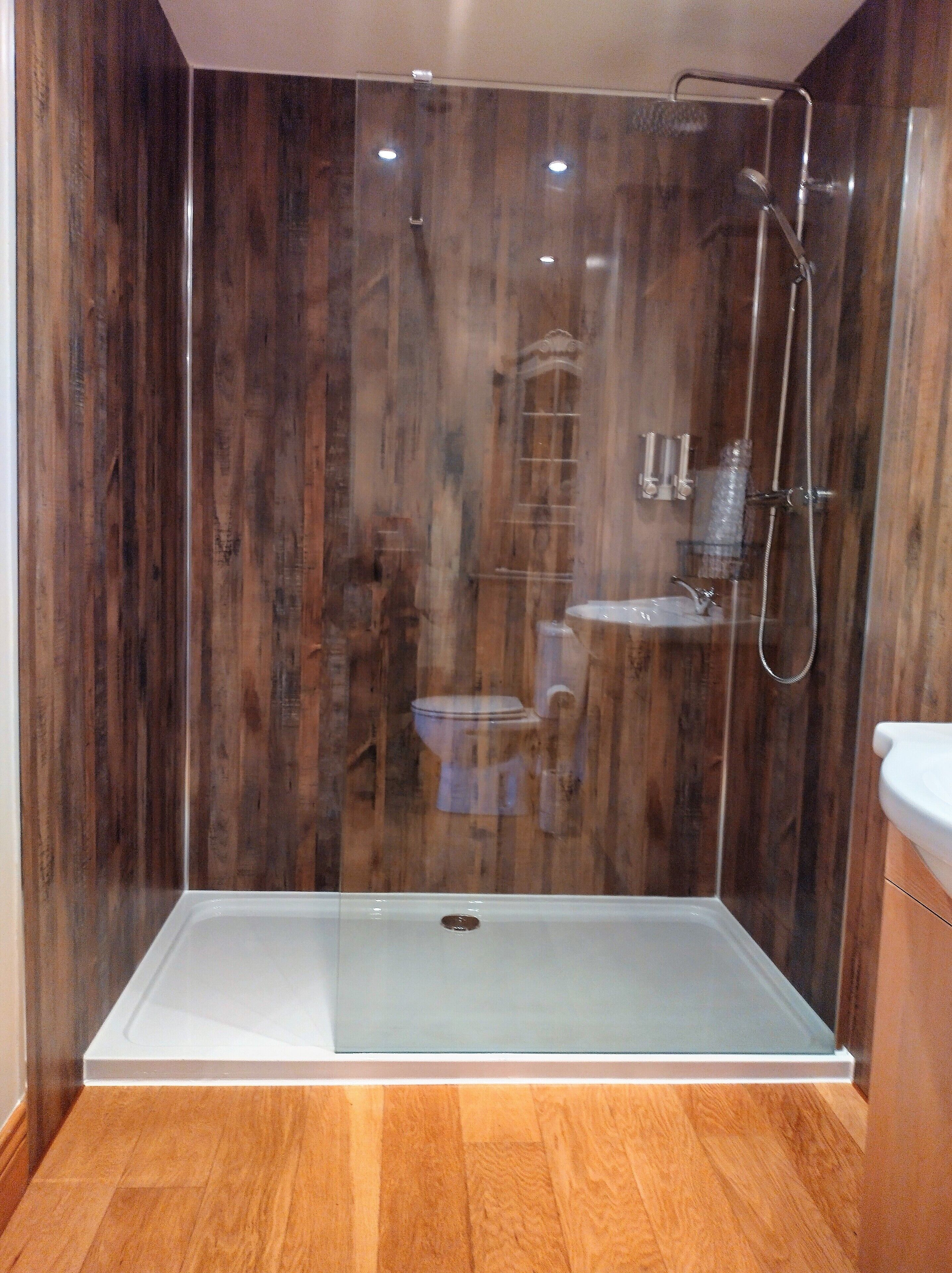 A glass-enclosed shower stall with a white sink and toilet, wooden paneling on the walls, and a wooden floor.