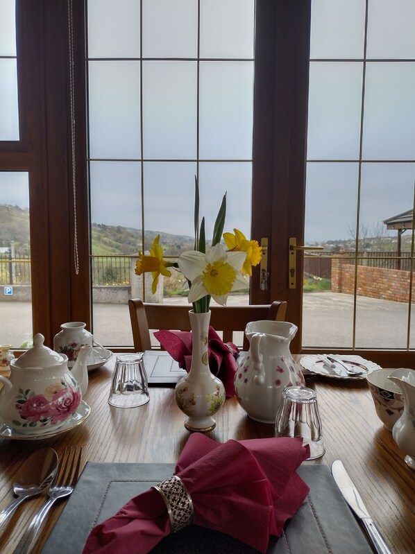 A dining table set with a vase of flowers, teapots, cups, and cutlery. - Glenn Eireann House (Ballymena)