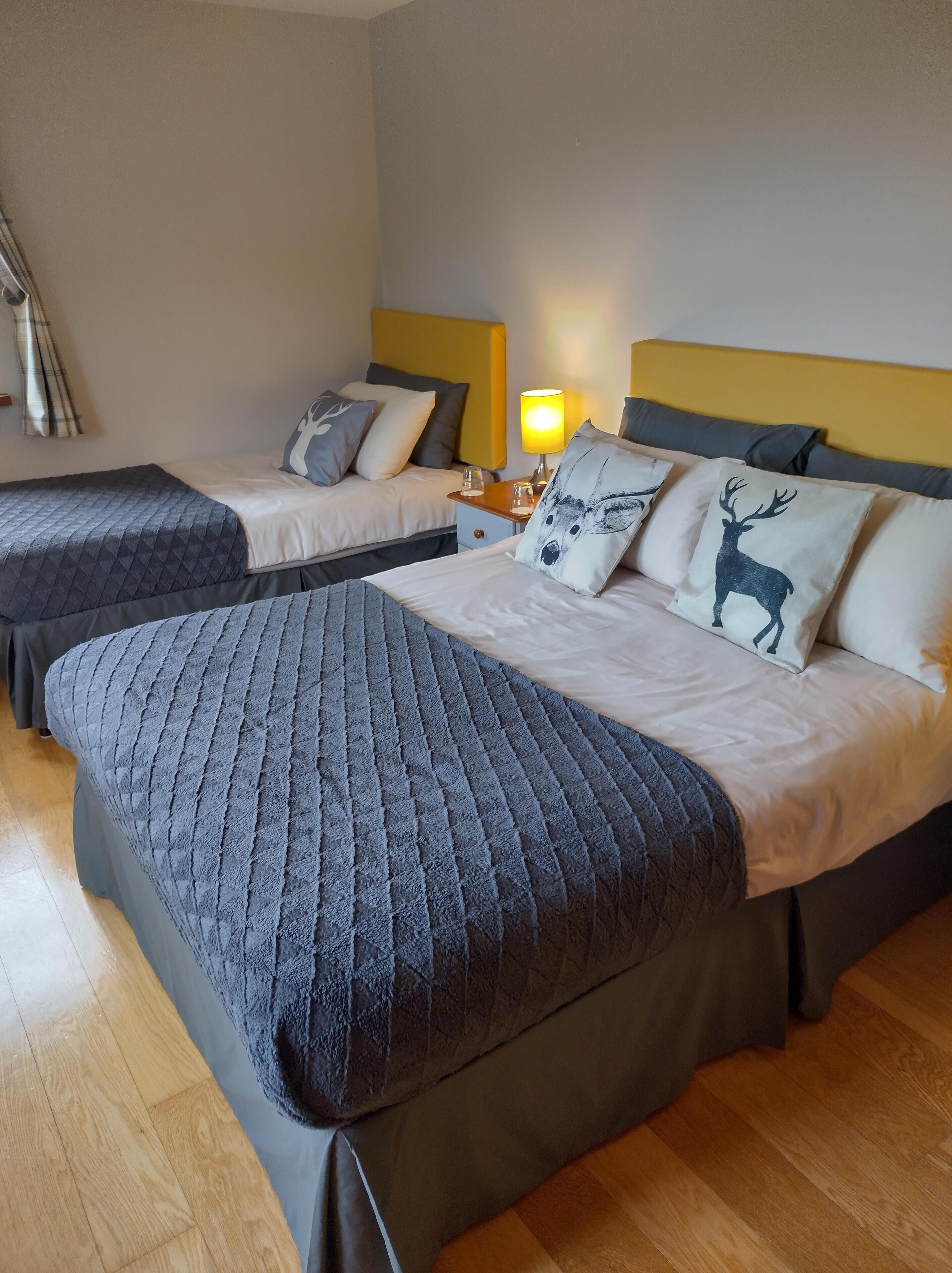 Habitación de hotel con dos camas, cada una con colcha gris acolchada y almohadas a juego. Entre las camas, una mesita de noche con una lámpara.