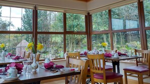 Un comedor con mesas y sillas de madera, amplias ventanas con vista a la vegetación y un candelabro.