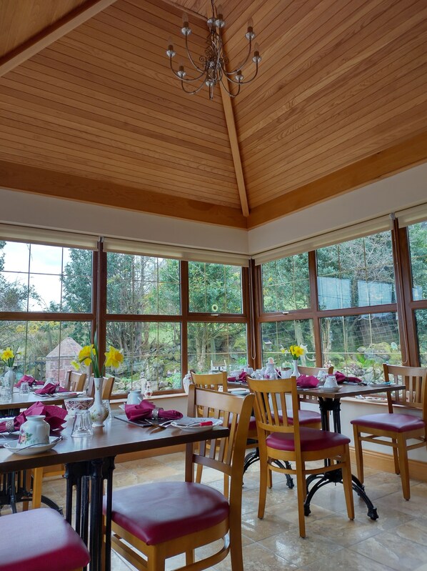 A dining area with wooden tables and chairs, large windows with a view of greenery, and a chandelier. - Glenn Eireann House (Ballymena)