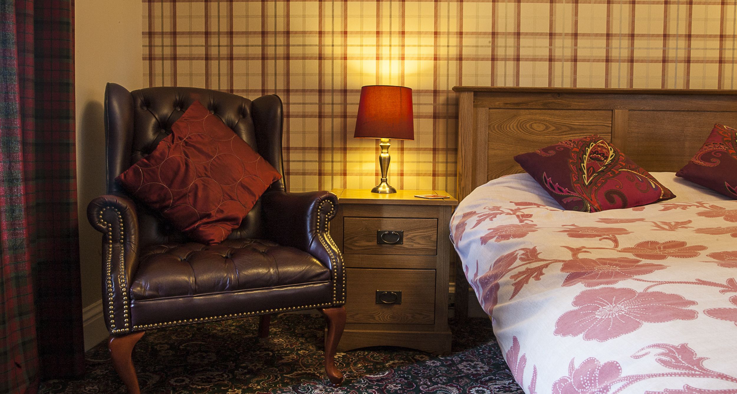 Habitación de hotel con un sillón de cuero, una mesita de noche de madera con lámpara, cama con ropa de cama floral y pared con estampado de cuadros.