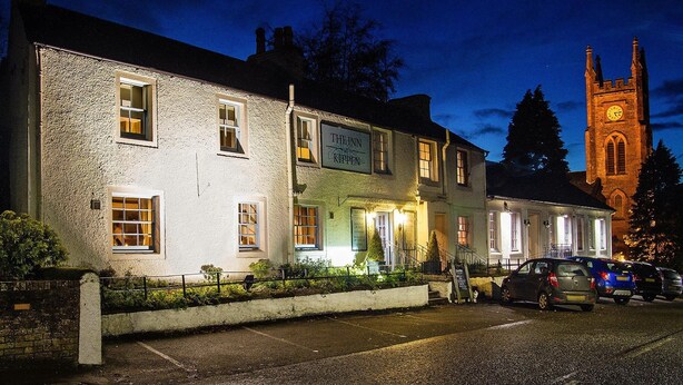 A traditional stone building with a sign reading "The Inn Kitchen" at night, parked cars, and a church tower in the background. - The Inn at Kippen (Stirling)