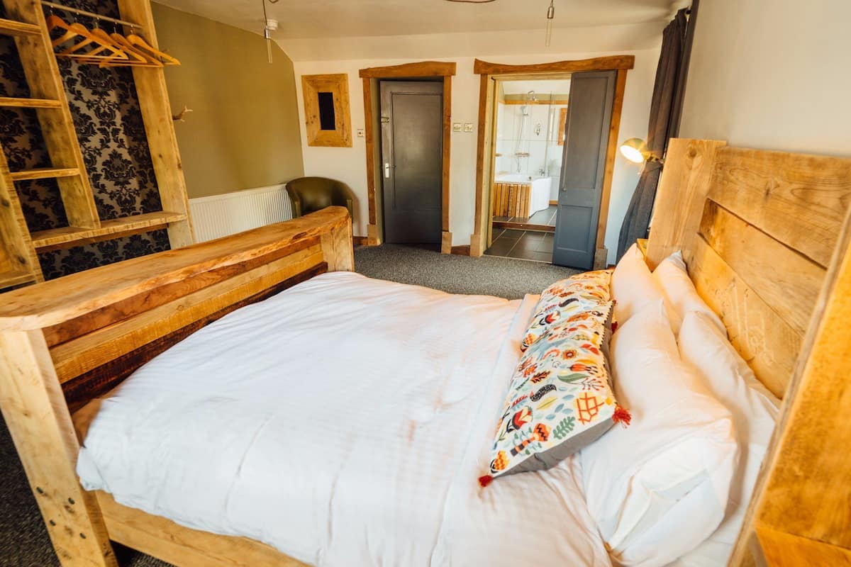 A wooden bed with a white mattress and colorful pillow, a wooden headboard, and a wooden ladder against a wall with patterned wallpaper.