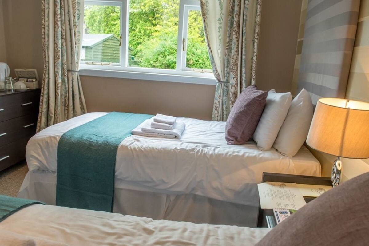 A bedroom with two beds, a window with curtains, a bedside table with a lamp, and a dresser.