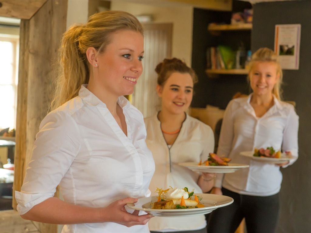 Trois personnes se trouvent dans une pièce dont le plafond est constitué de poutres en bois, et elles tiennent toutes dans leurs mains des assiettes remplies de nourriture.