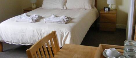 A neatly made bed with white linens, a wooden nightstand with a lamp, a wooden table with a tray of breakfast items, and a chair.
