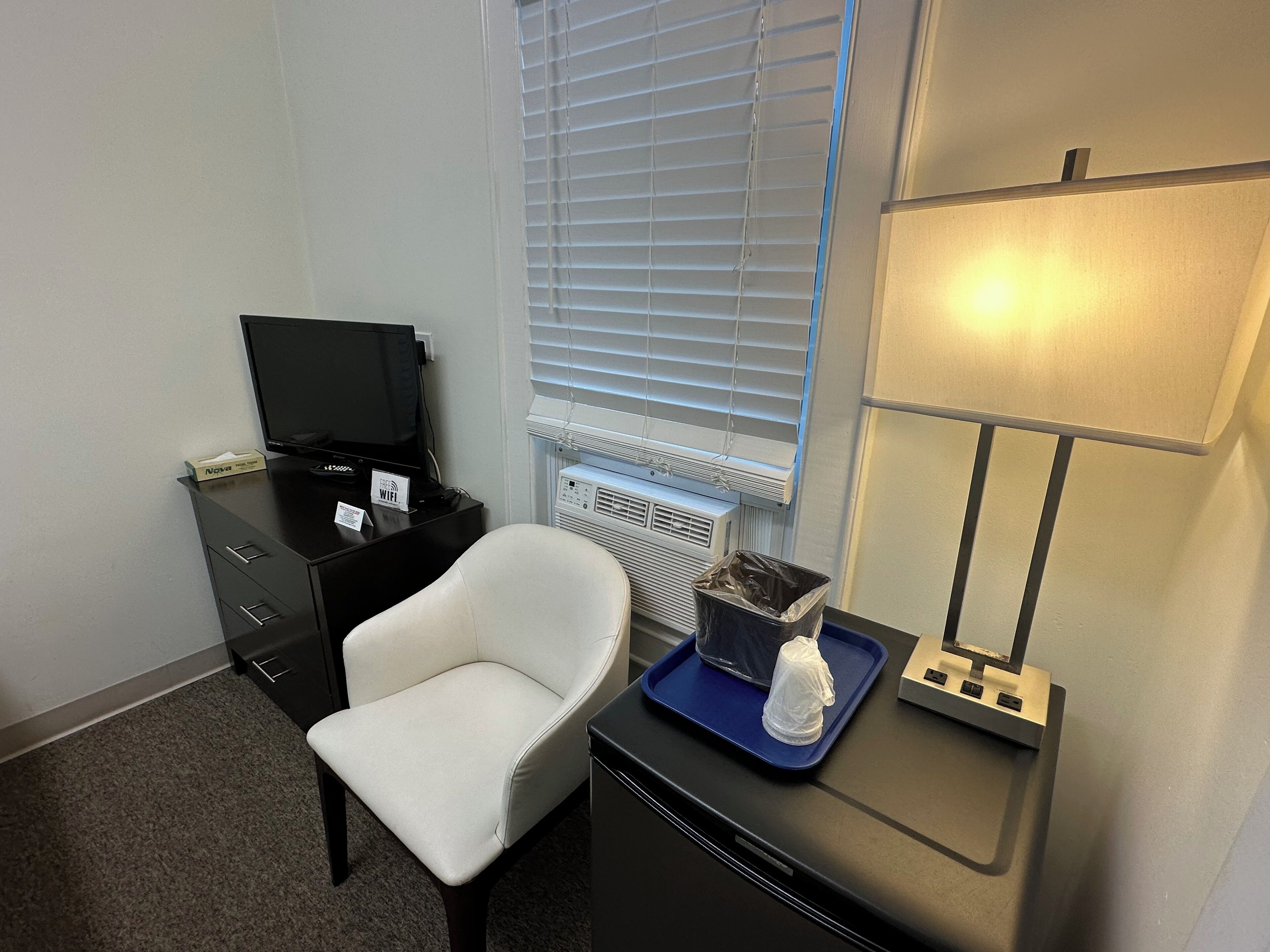 A hotel room with a desk, a chair, a television, a lamp, and a window with blinds.