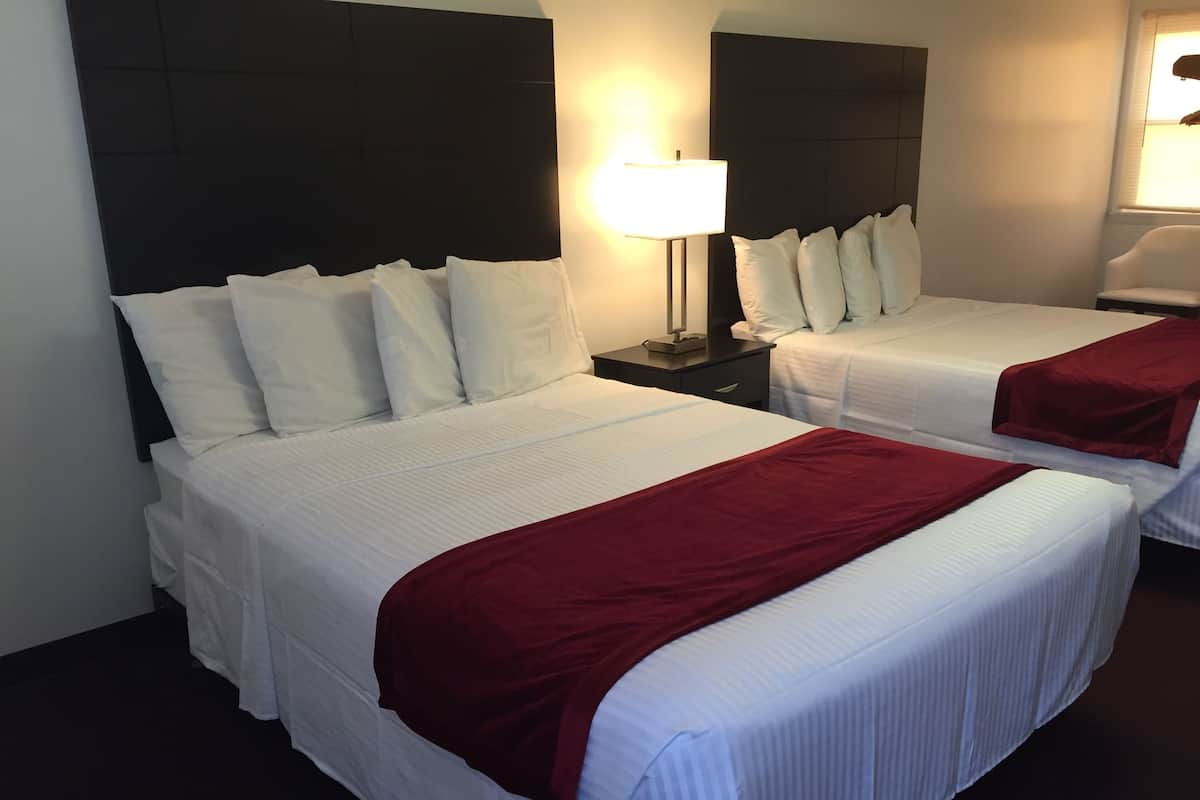 Habitación de hotel con dos camas, cada una con ropa de cama blanca y una manta roja. Entre las camas, una mesita de noche con una lámpara.