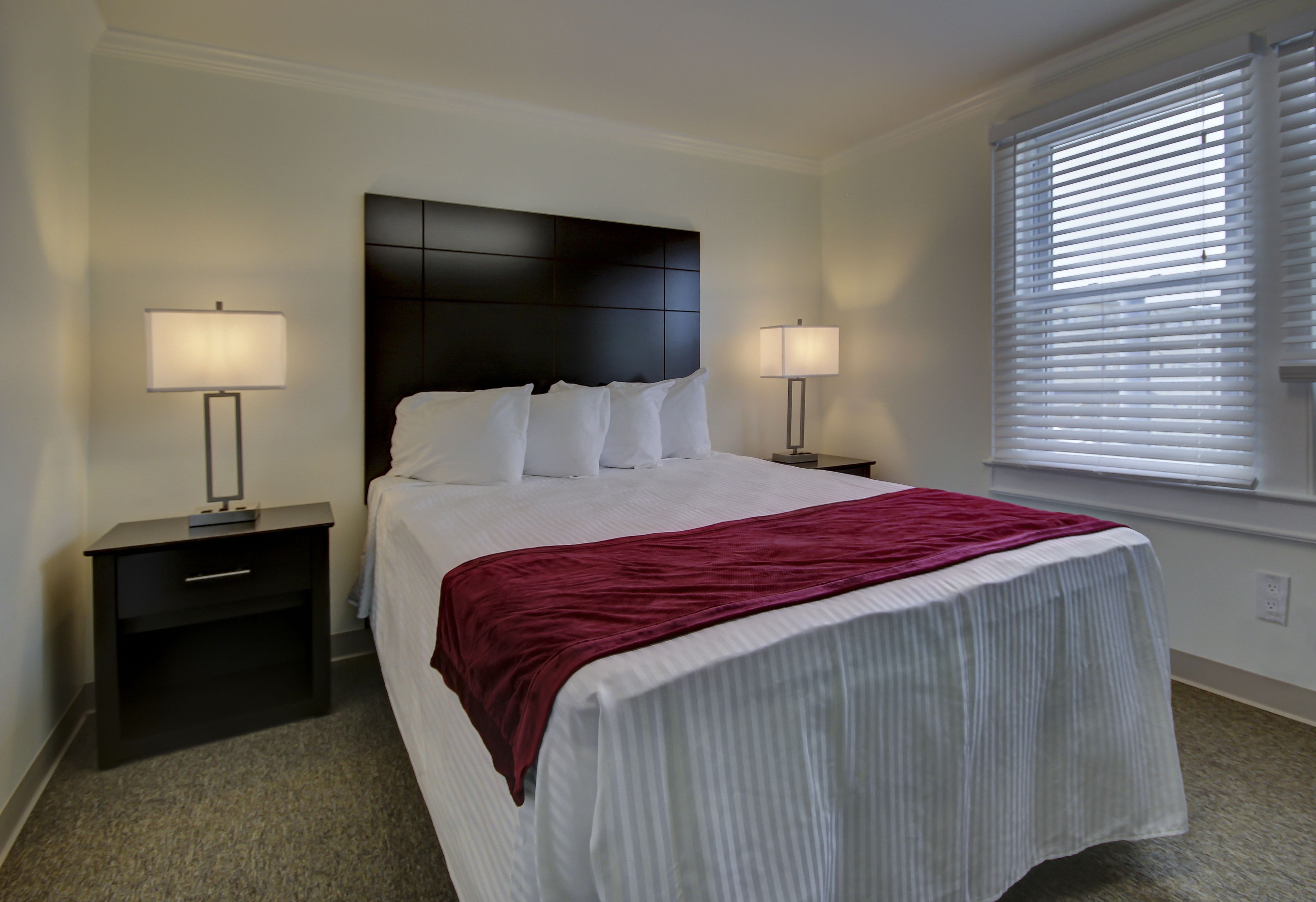 A neatly made bed with white linens and a red blanket, flanked by two bedside tables with lamps, in a room with a large window with blinds.