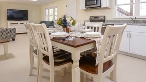 Un comedor con una mesa dispuesta para cuatro personas, sillas blancas, un televisor al fondo y una cocina con armarios blancos y un microondas.
