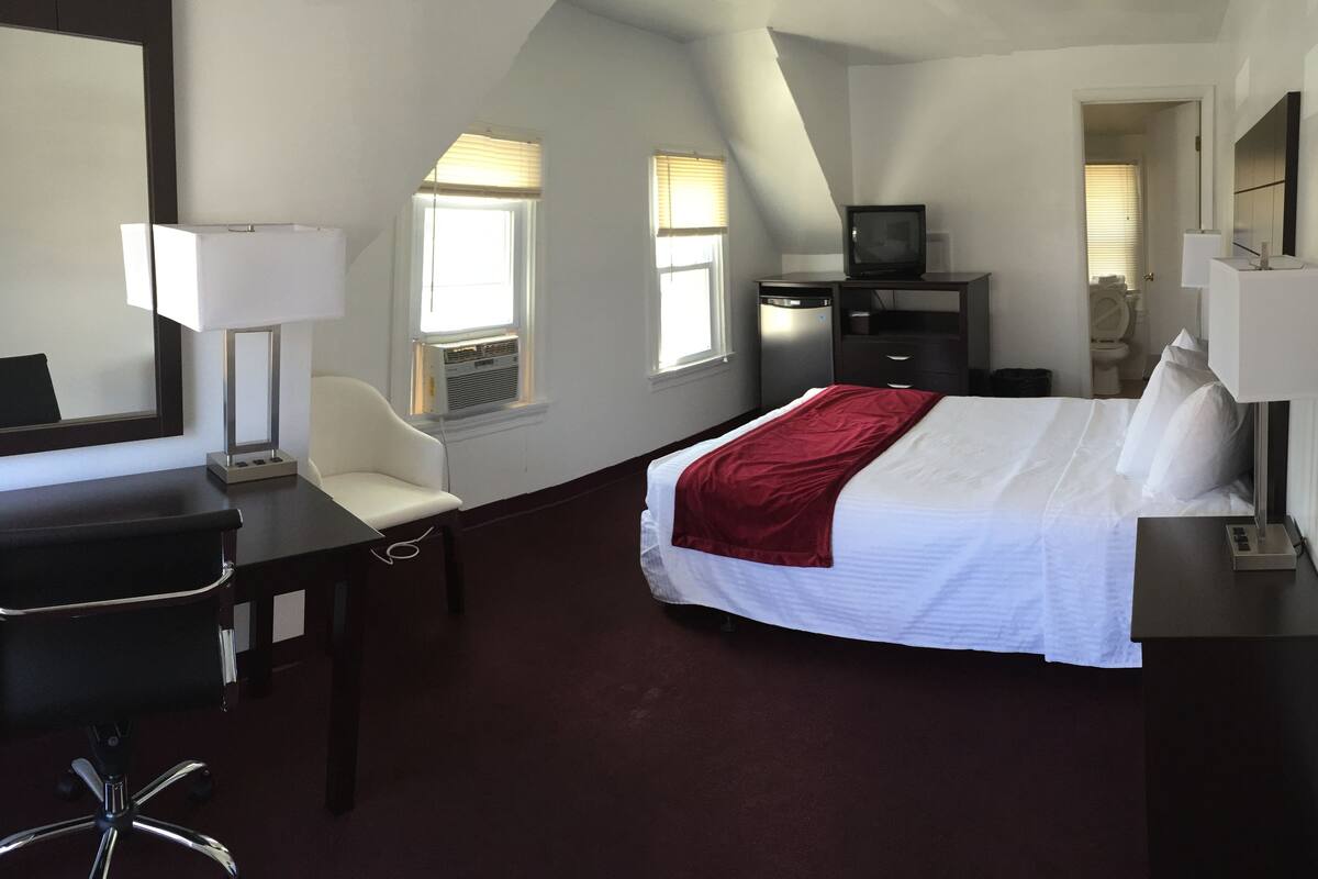 Habitación de hotel con cama, escritorio con silla, televisión y ventana con persianas.