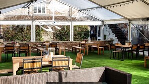A covered outdoor dining area with tables and chairs, a grassy lawn, and a stone building in the background. - The Bear and Swan (Bristol)
