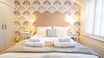 Une chambre d’hôtel comprenant un lit, deux tables de chevet, une commode, une fenêtre avec des stores et un mur orné d’un motif représentant un arbre.