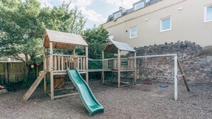 A wooden play structure with a slide and climbing area, surrounded by a stone wall and trees. - The Bear and Swan (Bristol)