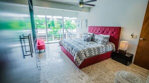 A bedroom with a red bed, a balcony with a view of greenery, and a ceiling fan.