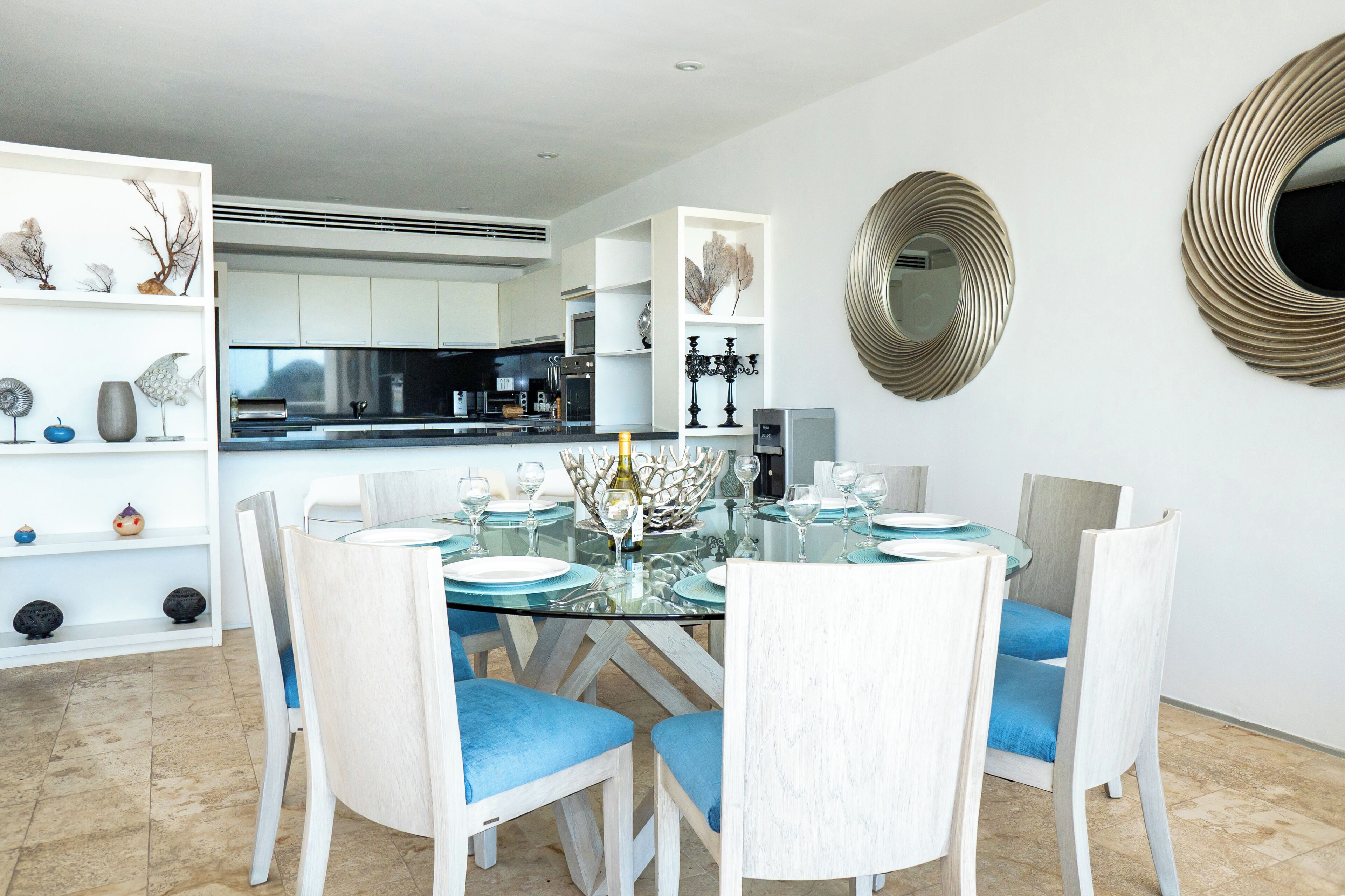 A modern dining area with a glass table, white chairs, and a kitchen in the background.