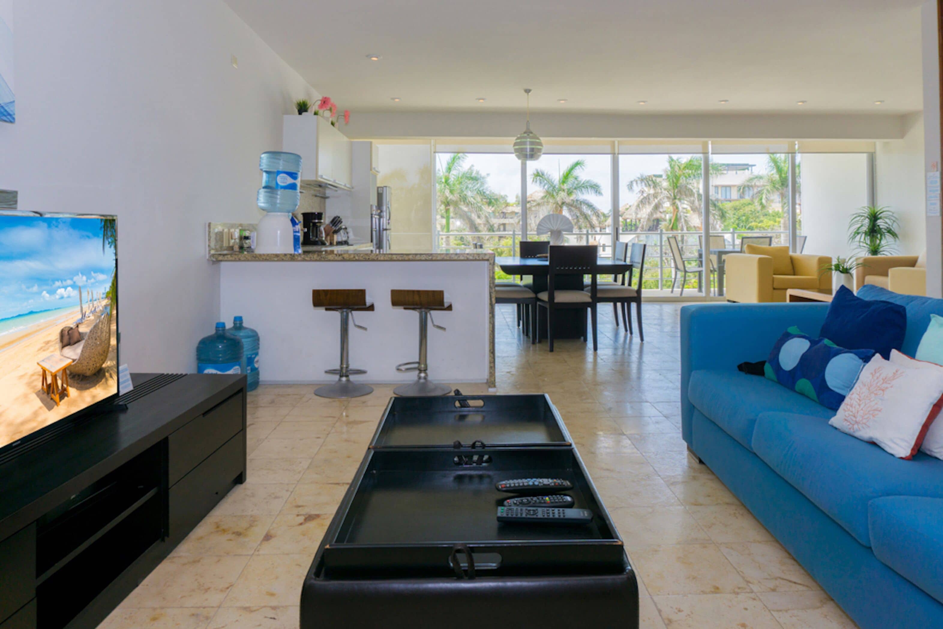 Ein modernes Wohnzimmer mit Fernseher, Couchtisch mit Fernbedienungen, einem blauen Sofa und einem Essbereich mit Stühlen und Tisch.
