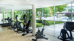 Een sportschool met diverse trainingsapparaten, een glazen wand en uitzicht op een zwembad en een terras.