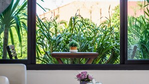 A room with a window overlooking a garden, a small wooden table with a potted plant, and a chair. - Baan Armeen Cottage (Choeng Thale)