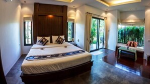 A modern bedroom with a large bed, a seating area with a table, and a view of greenery through the windows. - Baan Armeen Cottage (Choeng Thale)