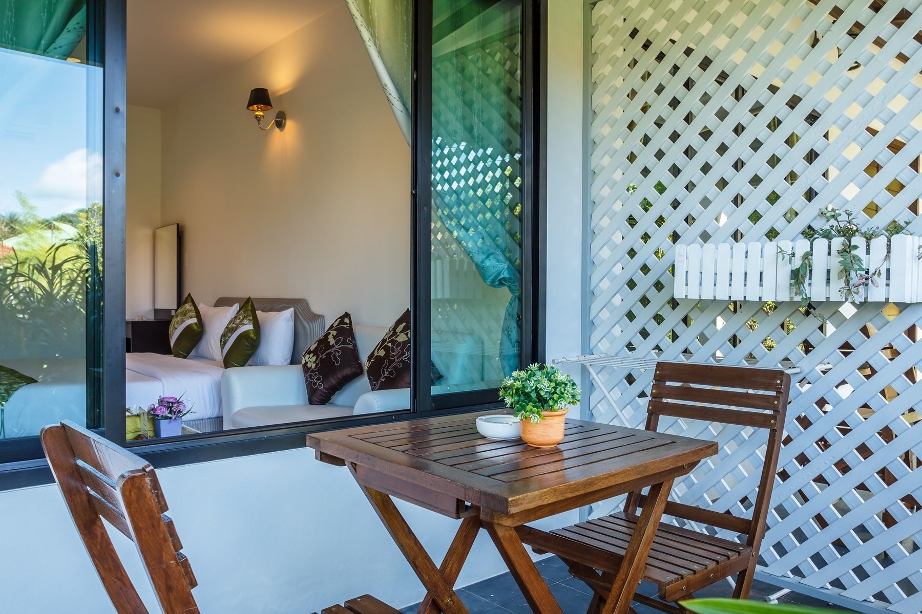 A wooden table and chairs on a balcony with a lattice wall and a window overlooking a room with a sofa and pillows.