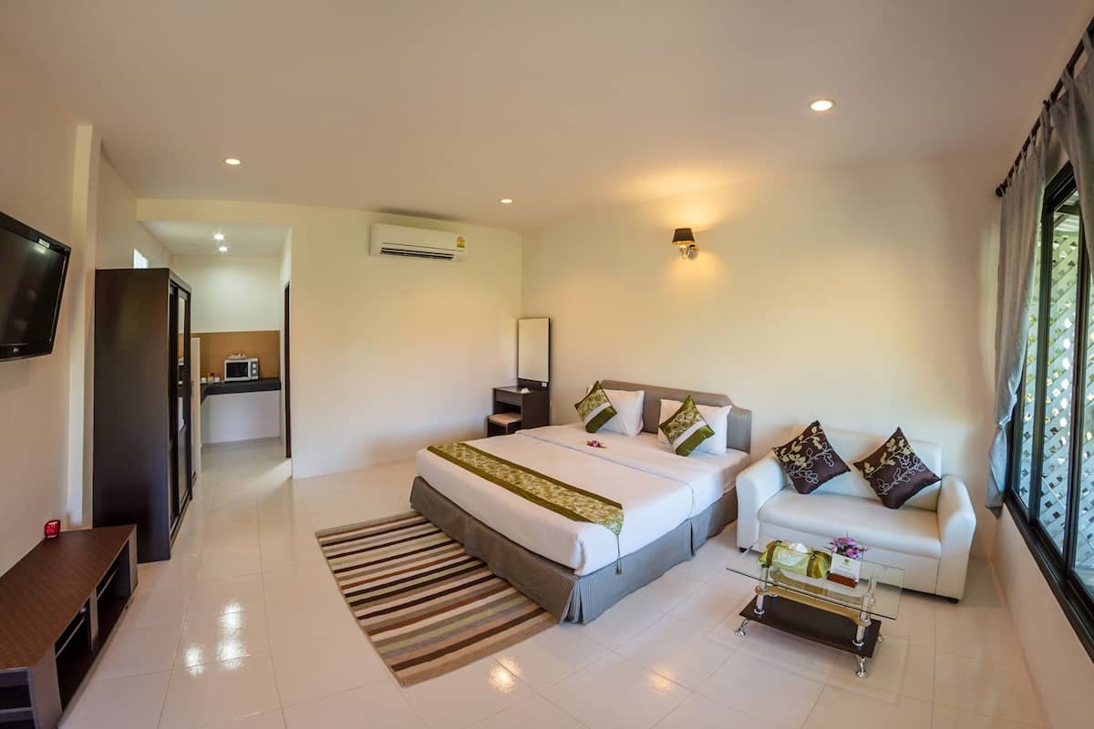 Habitación de hotel con cama, sofá, mesa de centro de vidrio, televisor y una kitchenette al fondo.