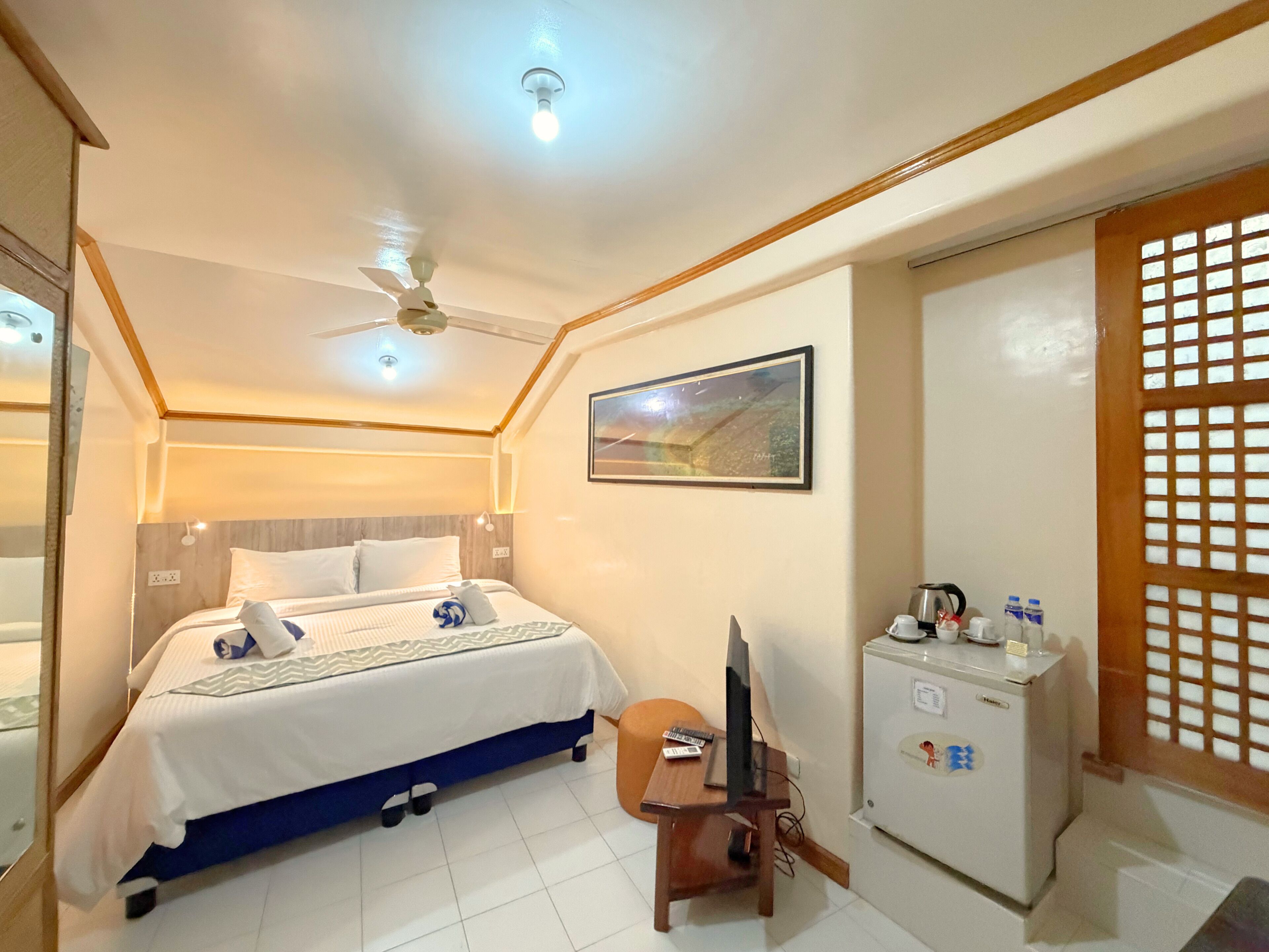 A hotel room with a bed, a desk with a computer, a refrigerator, and a ceiling fan.