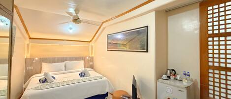 A hotel room with a bed, a desk with a computer, a refrigerator, and a ceiling fan.