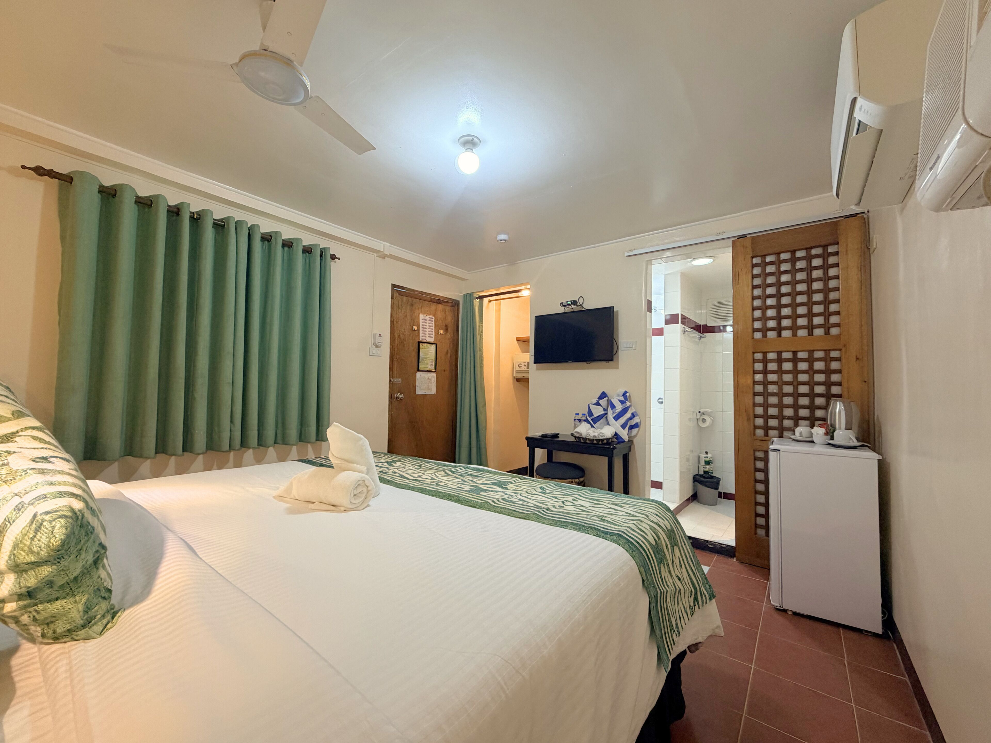 A hotel room with a bed, a ceiling fan, a television, a small table with a chair, and a refrigerator.