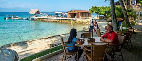 A seaside restaurant with outdoor seating, a view of the ocean, and a dock with boats.