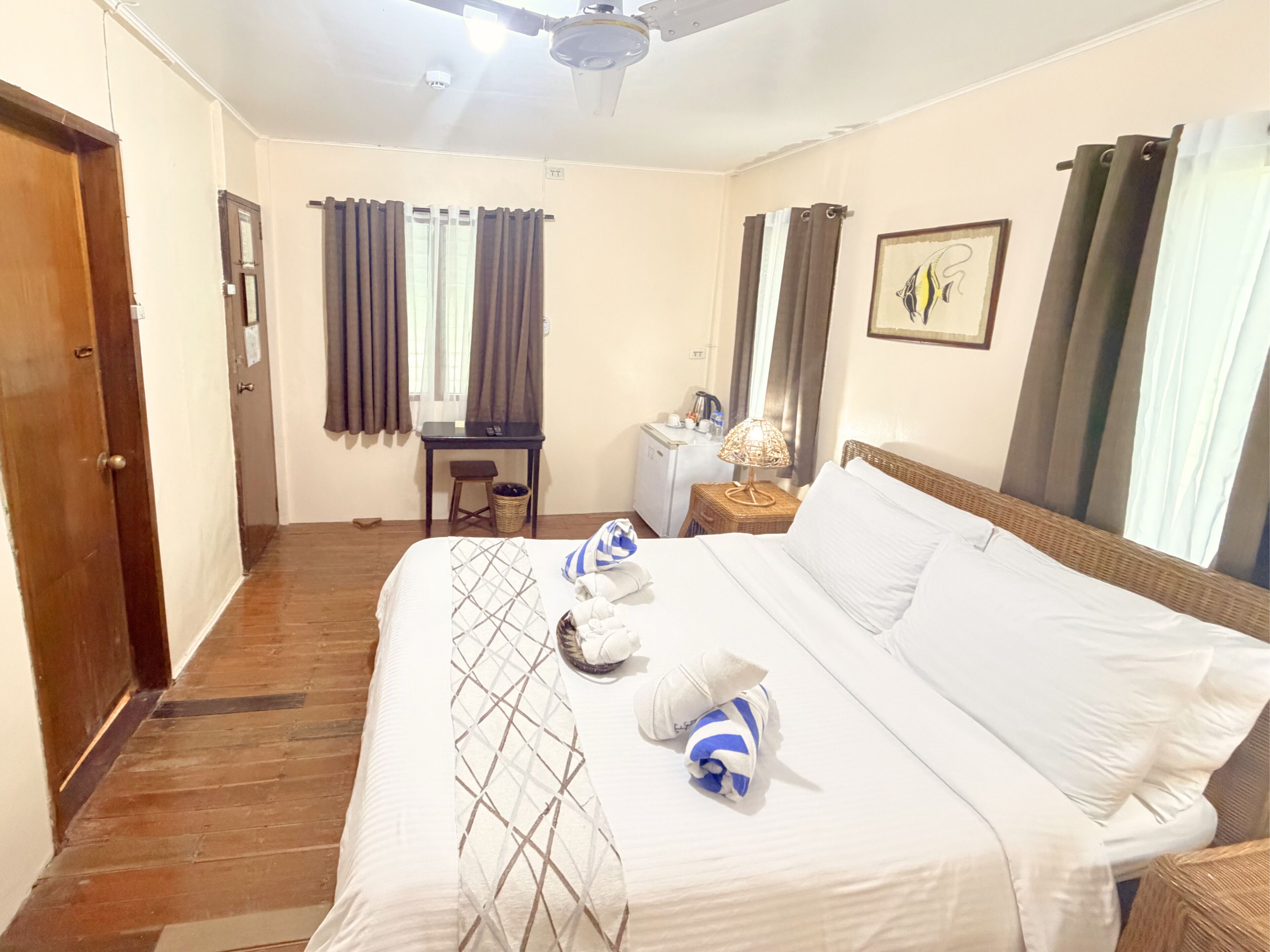 Habitación de hotel con cama, escritorio, silla, mesita de noche con lámpara, cesta y televisor de pared.
