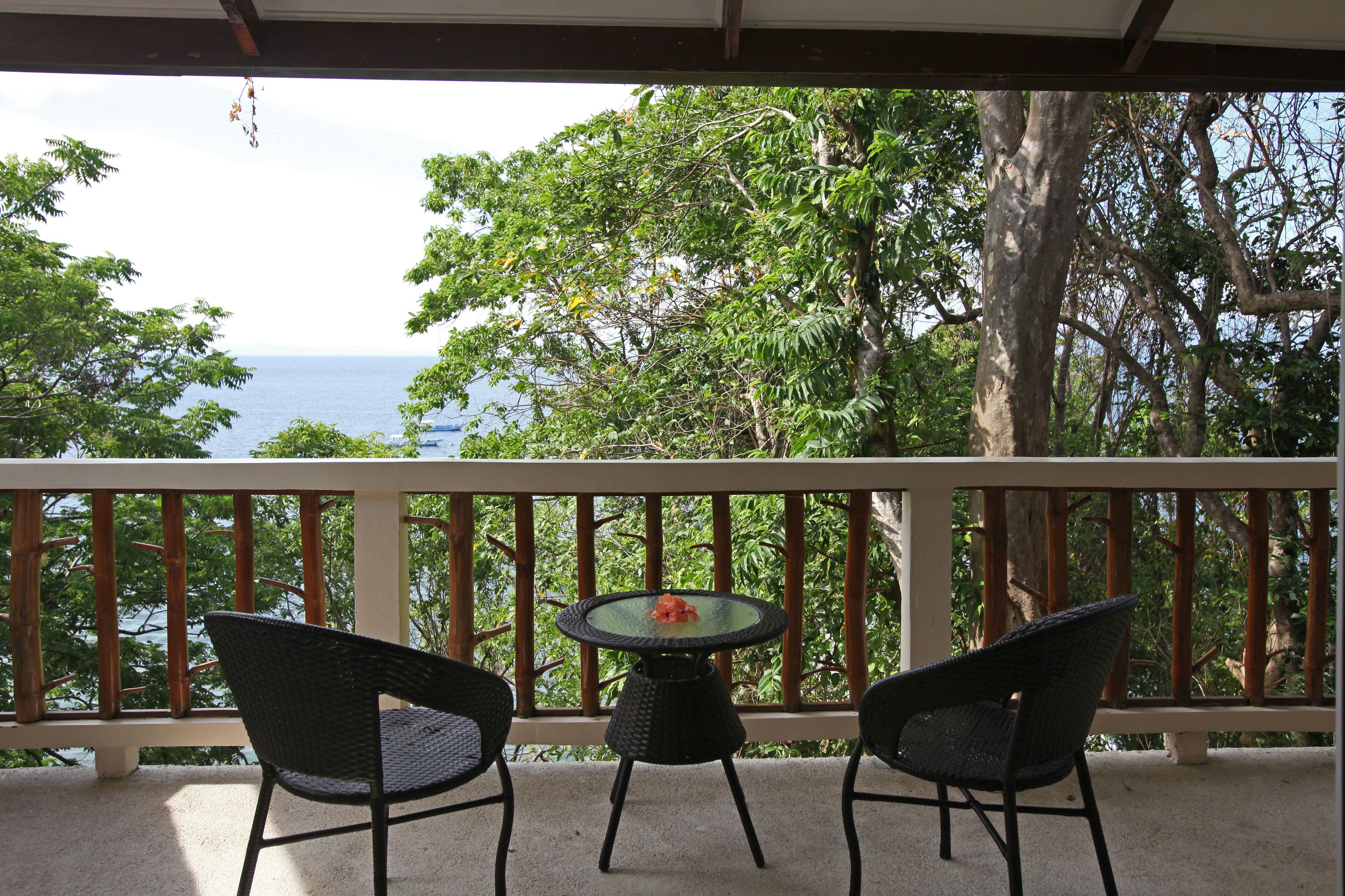 A balcony with two wicker chairs and a small round table, overlooking a body of water and lush greenery.