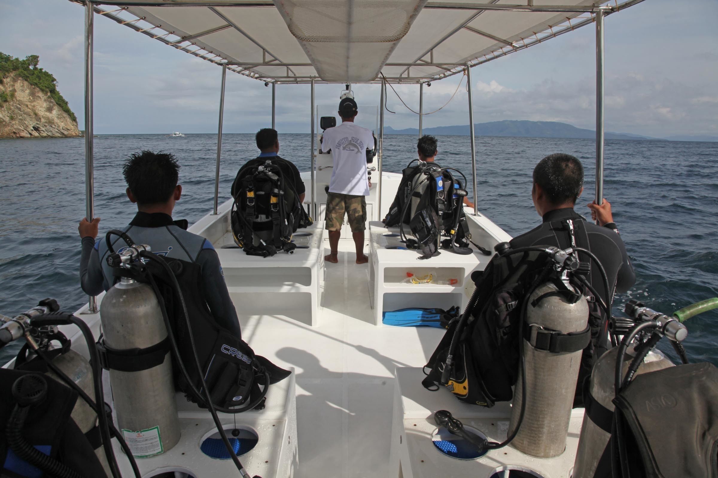 Un bateau avec trois plongeurs équipés de matériel de plongée sous-marine, une bâche blanche et une côte rocheuse en arrière-plan.