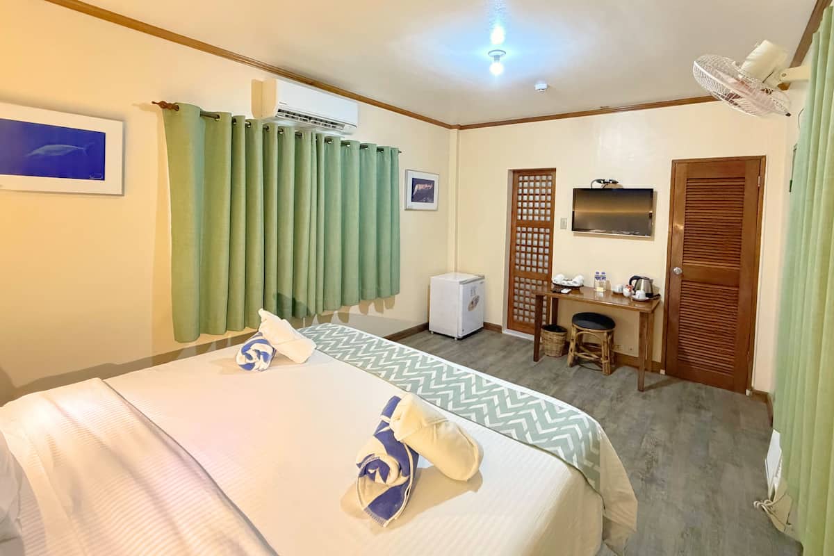 A hotel room with a bed, a desk, a chair, a small refrigerator, a fan, and a door.