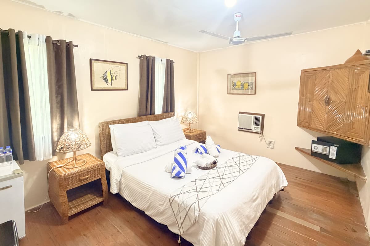 A hotel room with a bed, bedside tables, a ceiling fan, a wall-mounted air conditioner, and a wooden cabinet.